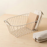 A white wire storage basket with rope handles sits on a light wood table, holding a beige notebook and a canvas pouch, with another matching pouch in the foreground.