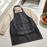 A black waterproof apron laid flat over a beige long-sleeve shirt on a kitchen counter, with water droplets shown on the front pocket to demonstrate water resistance. A sink and a bowl of fruit and tomatoes are visible in the background.