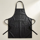 A black waterproof apron with a neck strap, waist ties, and a front pocket, shown flat against a light beige background with water droplets on the surface to demonstrate water resistance.