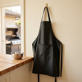 A black apron with a front pocket hangs from a wooden hook on a beige wall in a kitchen setting, with a wooden countertop and a stainless steel bowl nearby.