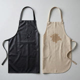 Two aprons laid flat on a light gray background: one black apron with a small dark stain graphic on the chest pocket area, and one beige linen-style apron with a brown stain graphic on the chest area. Both aprons are shown side by side.