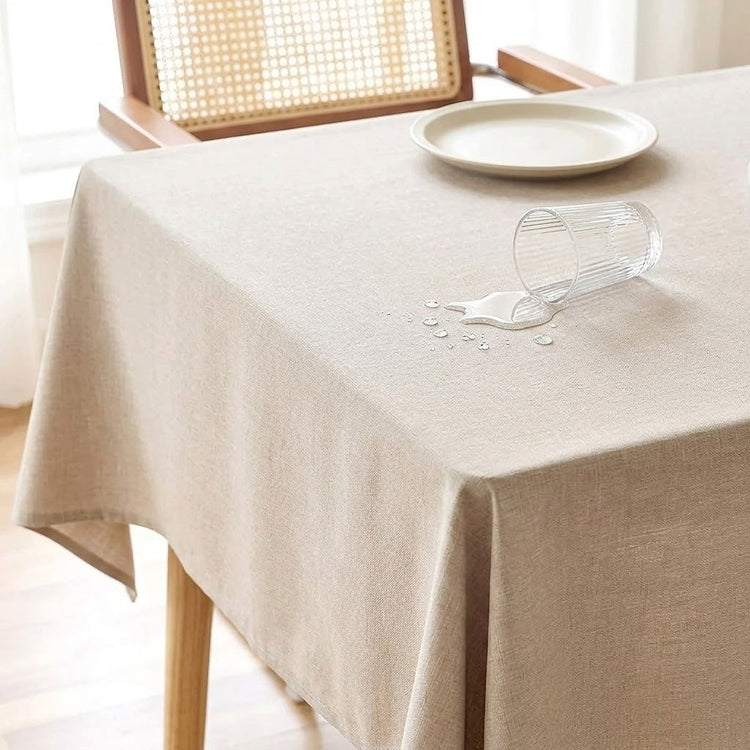A beige linen tablecloth on a wooden table, with a clear glass tipped over to show water droplets being absorbed by the fabric. A white plate and a woven-back chair are visible in the background.