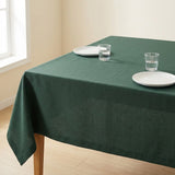 A dark green tablecloth draped over a rectangular wooden table in a bright room, styled with two white plates and two clear drinking glasses.