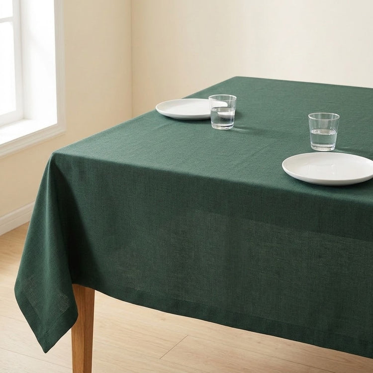 A dark green tablecloth draped over a rectangular wooden table in a bright room, styled with two white plates and two clear drinking glasses.