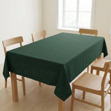 A dark green tablecloth draped over a rectangular wooden dining table with four light wood chairs in a bright, minimalist room.