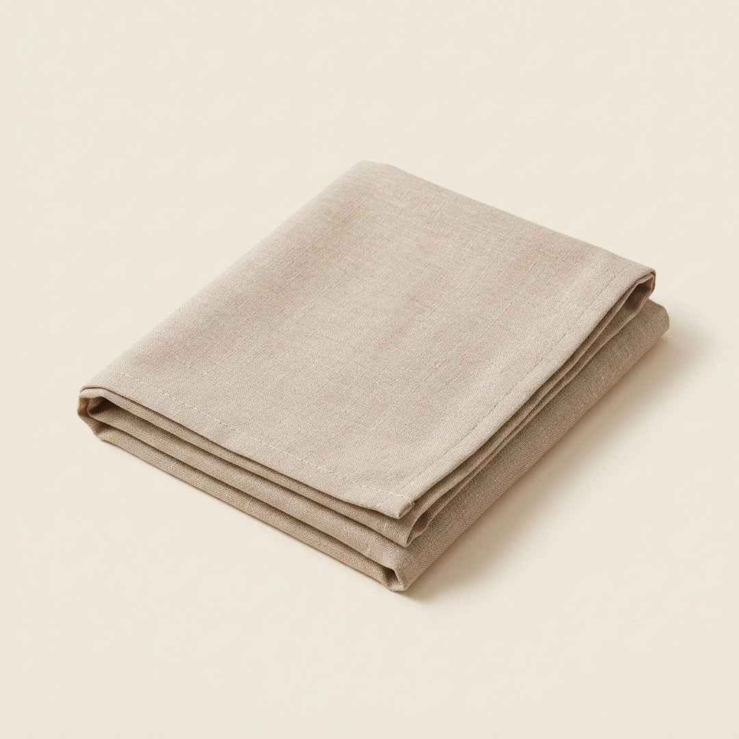 A folded linen tablecloth or napkin in a light taupe-beige color, displayed on a clean, solid cream background.