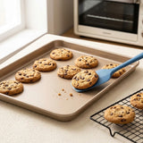 A beige silicone spatula with a blue handle is lifting a chocolate chip cookie from a beige baking sheet, with other cookies on a cooling rack in a bright kitchen setting.