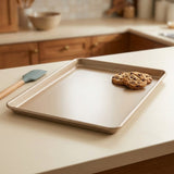 A rectangular metal baking sheet with raised edges sits on a light countertop in a kitchen setting. Two chocolate chip cookies rest on one corner of the tray, and a blue silicone spatula with a wooden handle lies nearby.