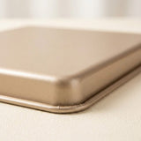 Close-up of a rectangular metallic tray or pan with rounded corners, shown on a light beige surface. The product has a warm champagne-gold finish and is photographed in soft, diffused lighting.
