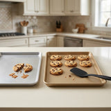 Two baking trays on a kitchen counter, one silver with a broken chocolate chip cookie and crumbs, and one beige with several whole chocolate chip cookies and a black spatula. The kitchen is softly blurred in the background.