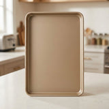 A rectangular non-stick baking sheet with rounded corners in a matte champagne gold finish, standing upright on a light marble kitchen countertop with a blurred kitchen background.