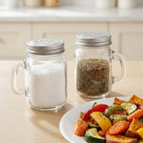 Two clear glass mason jar shakers with silver metal lids and handles sit on a light wood table, one filled with white salt and the other with dark pepper. A plate of roasted vegetables is partially visible in the foreground.