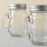 Two clear glass mason jar mugs with side handles and silver metal screw-on lids, shown on a light neutral background.