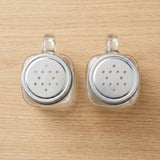 Two clear glass pepper shakers with silver metal shaker tops, displayed side-by-side on a light wood grain surface.