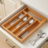 A bamboo drawer organizer tray arranged inside an open kitchen drawer, holding silver metal cutlery and chopsticks, with a few utensils placed outside on the drawer surface.