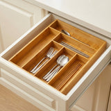 A bamboo drawer organizer tray arranged inside an open kitchen drawer, holding silver utensils and chopsticks. The tray has multiple compartments and is shown in a real kitchen setting with cream-colored cabinets.