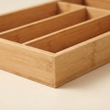 A close-up of a light natural bamboo organizer tray with multiple rectangular compartments, shown on a plain light beige background.