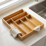 A rectangular bamboo kitchen organizer tray with multiple compartments, resting on a white countertop near a sink. Two small beige cloth napkins are tucked under the tray.