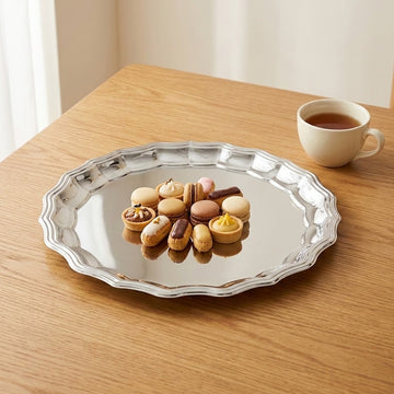 A round silver serving platter with a scalloped edge sits on a wooden table, holding assorted macarons and pastries. A white cup of tea is placed in the background on the right.