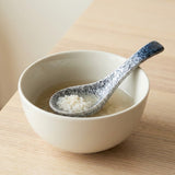 A ceramic bowl with a speckled blue and white spoon resting inside, containing a light broth or rice soup on a light wood tabletop.