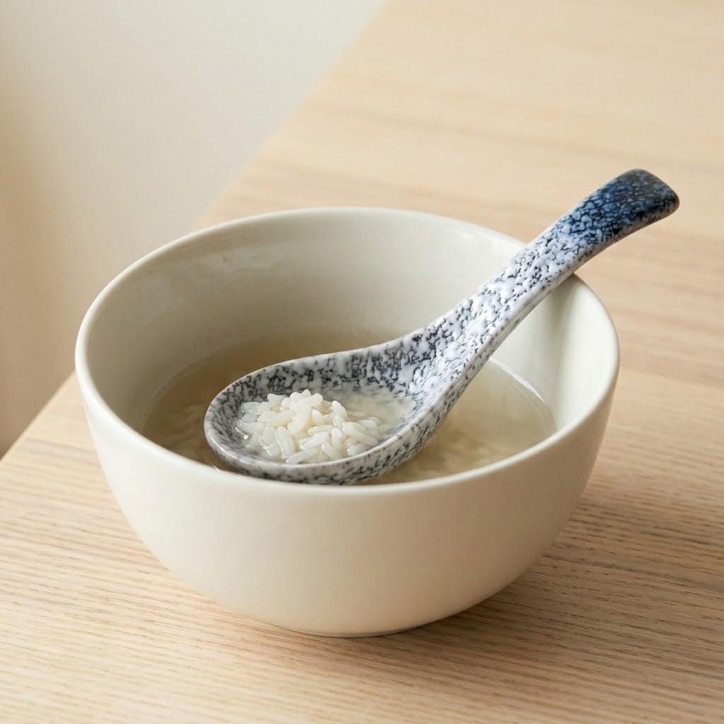 A ceramic bowl with a speckled blue and white spoon resting inside, containing a light broth or rice soup on a light wood tabletop.