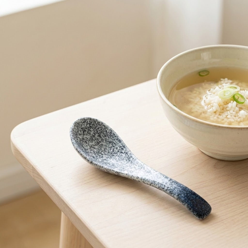 A speckled ceramic spoon with a dark blue gradient handle rests on a light wood table next to a bowl of miso soup with rice and green onions.