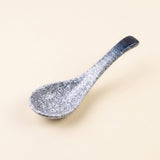 A ceramic spoon with a speckled blue and white glaze, featuring a dark blue handle, set against a solid light beige background.
