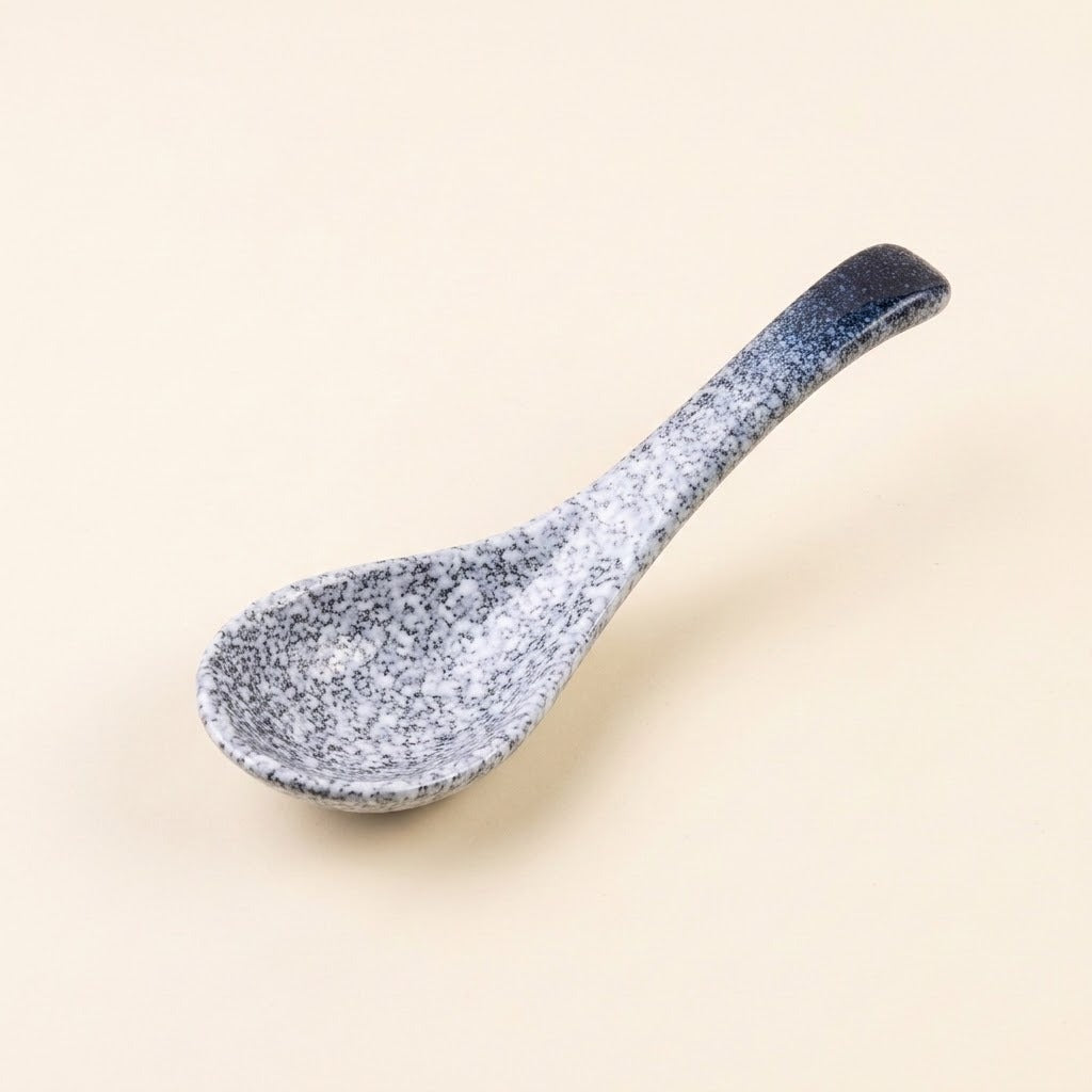 A ceramic spoon with a speckled blue and white glaze, featuring a dark blue handle, set against a solid light beige background.