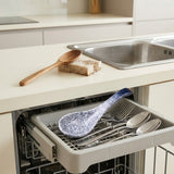 A dishwasher drawer is open in a bright kitchen, holding a speckled blue and white serving spoon and several stainless steel utensils. A wooden spoon and a sponge sit on the countertop beside a sink.