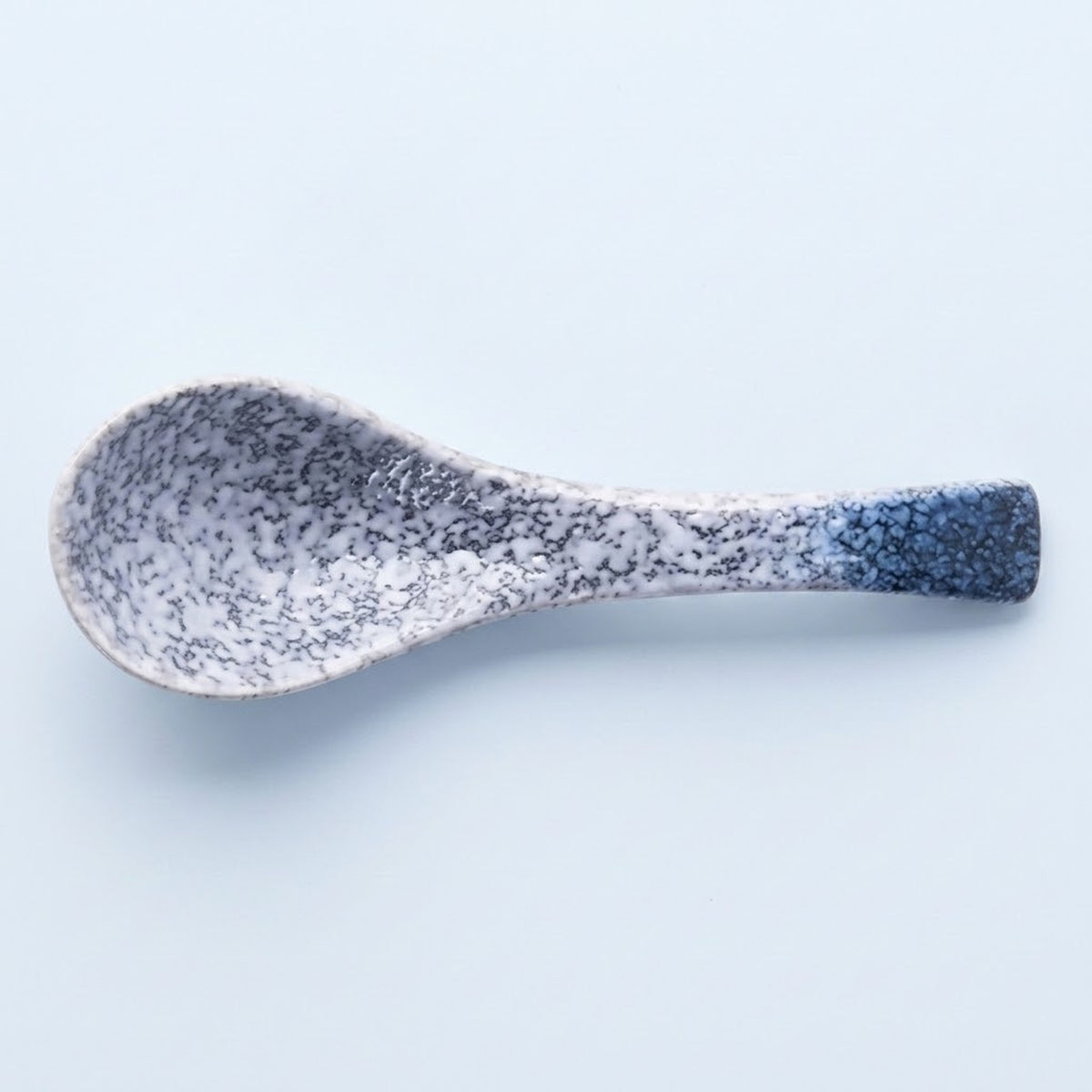 A ceramic spoon with a speckled glaze that transitions from white and gray to dark blue at the handle end, shown on a light blue background.