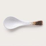A white ceramic spoon with fine brown speckles and a dark brown speckled handle, shown on a light beige background.