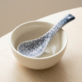 A ceramic bowl in warm off-white with a speckled blue-and-white spoon resting inside it, placed on a light wood surface against a soft beige background.
