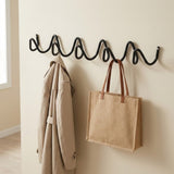A black wavy wall hook rack mounted on a beige wall, with a beige trench coat hanging on the left hook and a natural jute tote bag with brown handles hanging on the right.