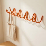 An orange spiral-shaped wall hook rack mounted on a cream-colored wall, with a beige canvas tote bag hanging from one of the hooks. A wooden table edge is visible in the bottom left corner.