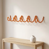 An orange wavy wall-mounted coat rack with hooks, shown above a light wood console table with a small white vase against a warm off-white wall.