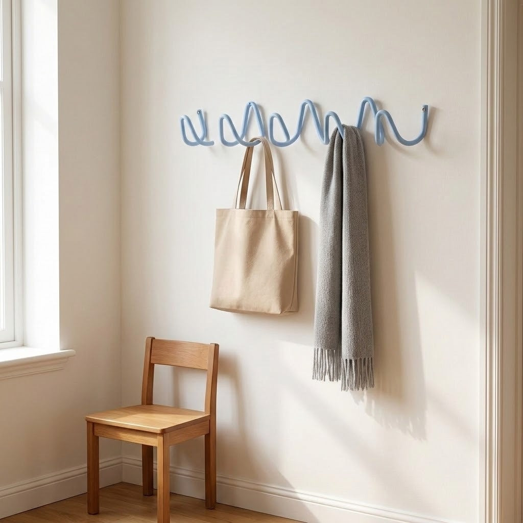 A blue wavy wall hook rack mounted on a cream-colored wall in a sunlit room, holding a beige tote bag and a gray scarf. A wooden chair sits below the rack.