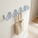 A light blue wavy wall hook rack mounted on a cream-colored wall, with a beige canvas tote bag hanging from one of the hooks in a bright indoor room.