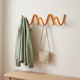 An orange wavy zig-zag wall coat rack mounted on a light beige wall, holding a sage green jacket and a beige woven shoulder bag, with a light wood bench visible at the bottom.