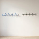 Two wall-mounted coat racks on a plain off-white wall above a light wood floor. The left rack is a wavy, openwork metal hook rack in light blue.