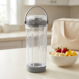 A clear ribbed travel tumbler with a gray lid and handle, standing on a kitchen counter next to a bowl of fruit and a cream tote bag.