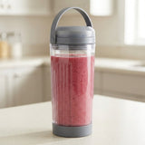 A clear ribbed smoothie bottle with a gray lid and handle, filled with a pink smoothie, sitting on a kitchen counter with a blurred kitchen background.