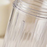 Close-up of a clear ribbed plastic blender jar cup with a threaded opening, shown against a softly blurred warm beige background.