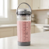 A clear plastic shaker bottle with a gray lid and handle, filled with a pink smoothie, standing on a light countertop with a blurred kitchen background.