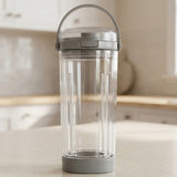 A clear plastic portable blender cup with a gray lid, carrying handle, and base, standing on a light countertop with a blurred kitchen background.