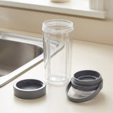 A clear plastic travel tumbler bottle with a gray lid and carrying loop, shown disassembled on a light beige countertop next to a stainless steel sink.