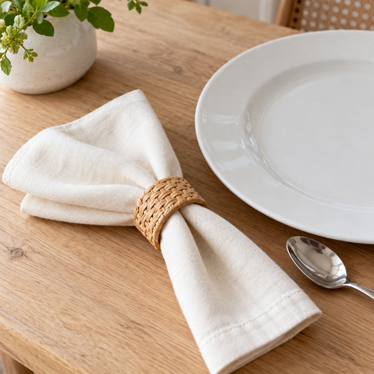 A natural tan rattan napkin ring beside a dinner plate and teaspoon shows its compact table scale.