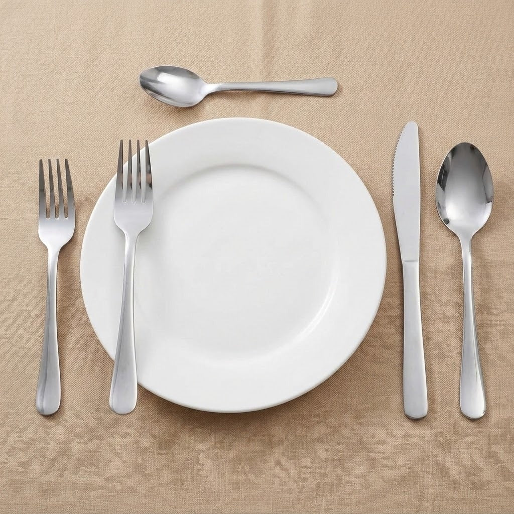 A place setting with a white round plate, two silver forks, a silver knife, and two silver spoons arranged on a beige fabric background.