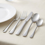 A set of polished stainless steel cutlery, including a fork, knife, and two spoons, arranged diagonally on a light beige textured tablecloth next to a white plate.