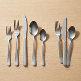 A flat set of stainless steel cutlery, including forks, knives, and spoons, arranged neatly on a light wood grain surface.
