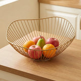 A gold-toned wire fruit bowl filled with apples and oranges, placed on a wooden countertop in a kitchen setting.